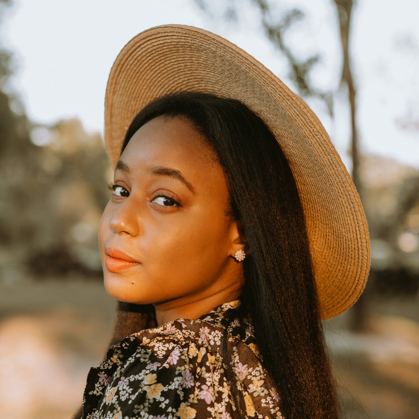 Chelsea, founder of Poppy+Lily wearing a straw hat in an outdoor setting with blurred trees and light in the background