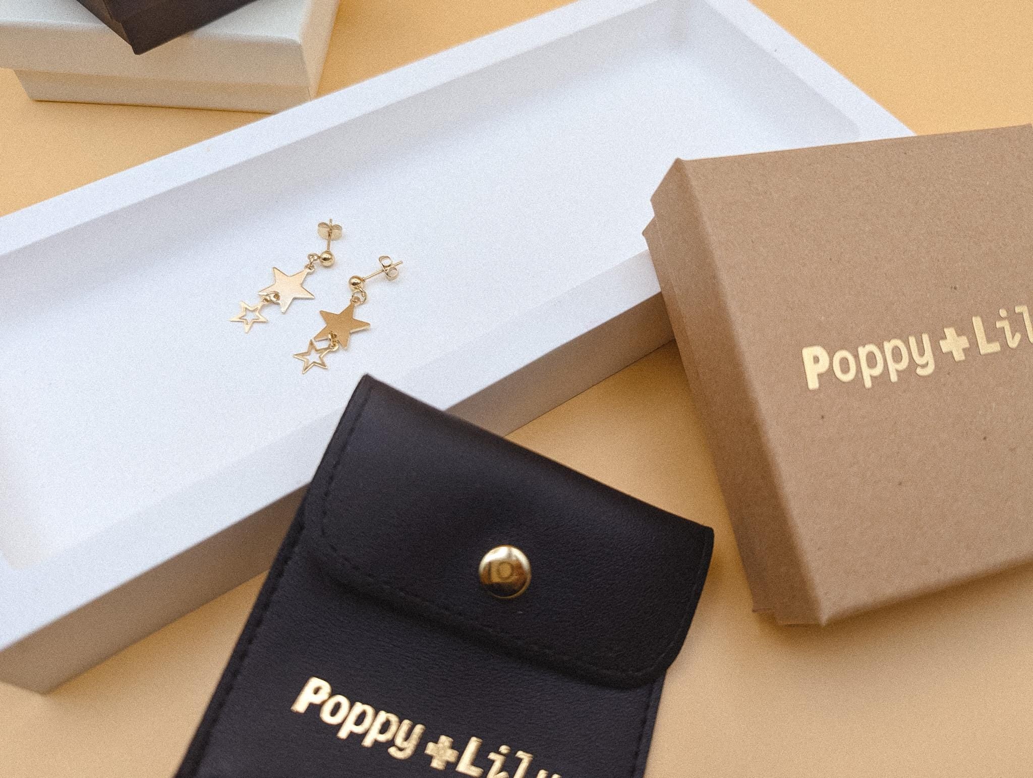 Gold star-shaped earrings in a jewelry pouch with 'Poppy + Lily' branding on a beige surface.