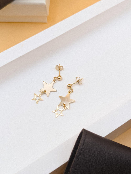 Gold star-shaped earrings on a white surface with a black box and yellow object in the background